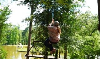 Person zip-lining over a forest with a river below, wearing a cap and holding onto the handle