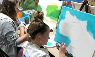 Two people painting side by side one on a landscape, the other outlining an animal on blue wall