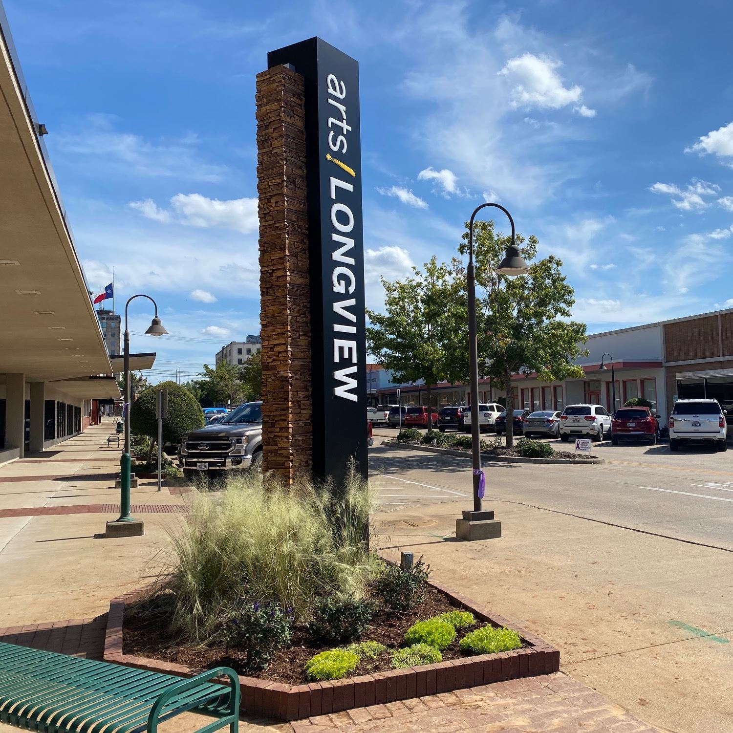 An 'arts! LONGVIEW' sign rises from a stone base near a planter and bench, set against Tyler S